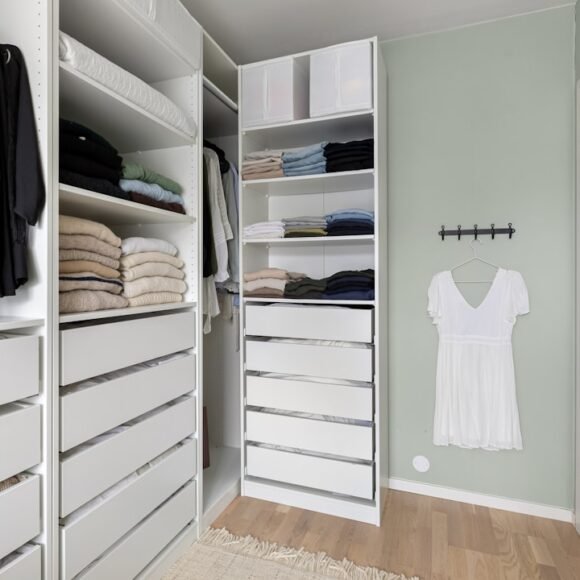 A small walk-in closet neatly organized with white drawers, hanging clothes, and labeled storage bins