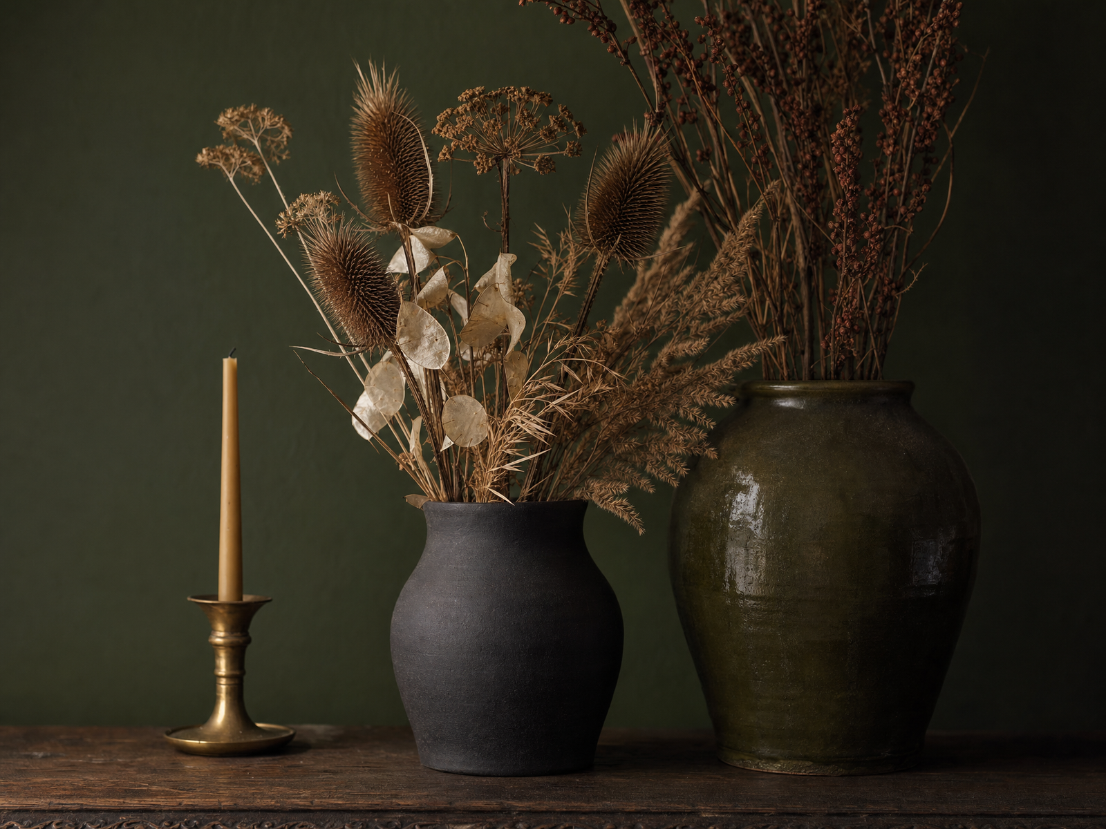 Dried seed heads and bleached grasses in matte charcoal and olive ceramic vessels beside a brass candlestick against a deep green wall