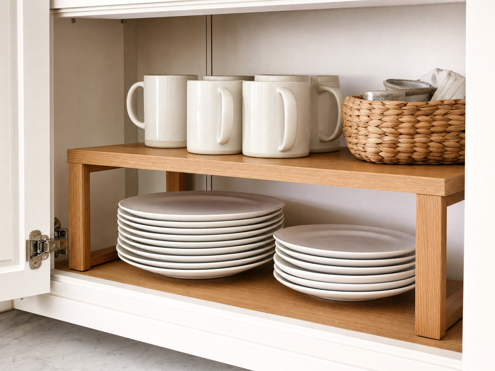Open kitchen cabinet fitted with a wooden two-tier shelf riser holding stacked white plates below and cream mugs above
