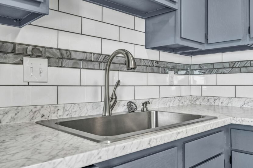 White subway tile backsplash above a marble look counter with a stainless steel sink and gray kitchen cabinets
